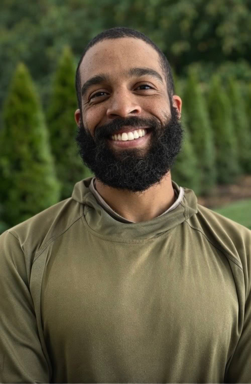 A man with a full beard and mustache smiles while standing outdoors in front of green trees, wearing an olive-green hoodie—a perfect look for someone working with Nyman Services in landscape construction.