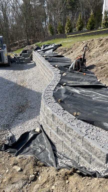 Curved retaining wall under construction with stacked concrete blocks, gravel base, black landscape fabric, and construction equipment visible on site—a showcase of quality landscape construction by Nyman Services.