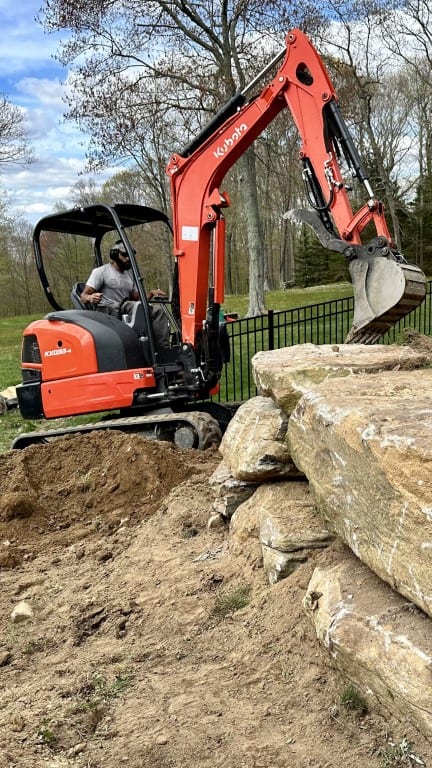 A person operates an orange Kubota mini excavator to move dirt and rocks near a stack of large stone slabs, showcasing landscape construction by Nyman Services in an outdoor area with trees and grass.