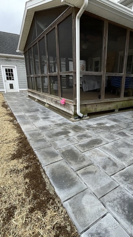 Concrete paver walkway bordering a screened porch, expertly crafted by Nyman Services. This landscape construction features a downspout and straw-covered ground along the edge for added functionality.