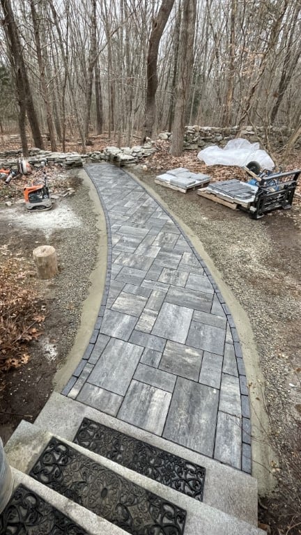A newly installed stone walkway with a herringbone pattern curves through a yard, showcasing expert landscape construction by Nyman Services. The path is bordered by unfinished landscaping, bare trees, and nearby construction materials.