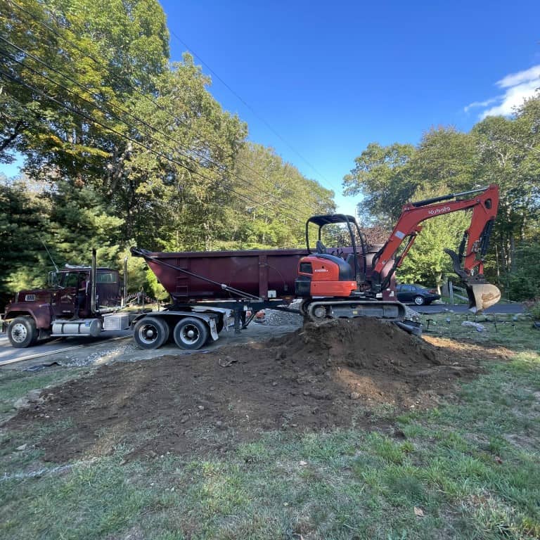 A small excavator from Nyman Services sits atop a dirt pile, loading soil into a large dumpster trailer attached to a parked truck—a scene of active landscape construction near a road.