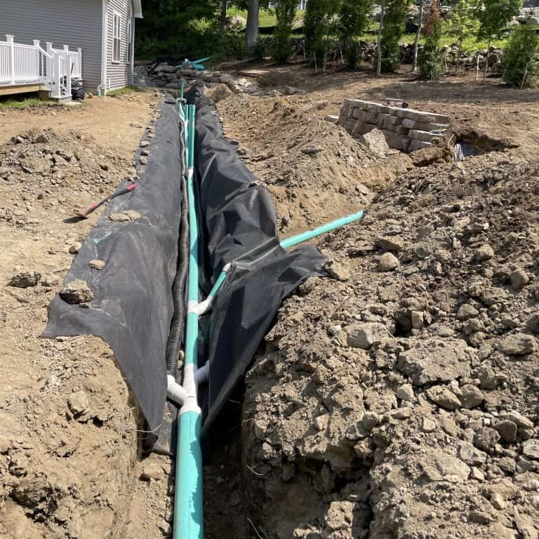 A trench with green pipes laid for drainage or utilities runs beside a house, surrounded by dirt piles and construction materials, showcasing nyman services' expertise in landscape construction.