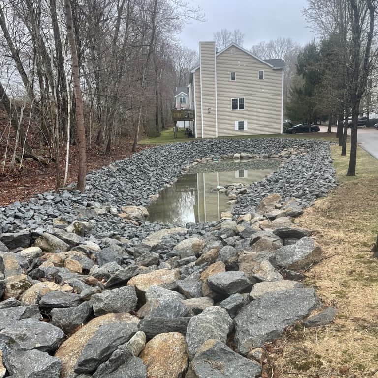 A retention pond lined with large gray rocks, a result of expert landscape construction by Nyman Services, is surrounded by trees and grass near a beige residential building on a cloudy day.