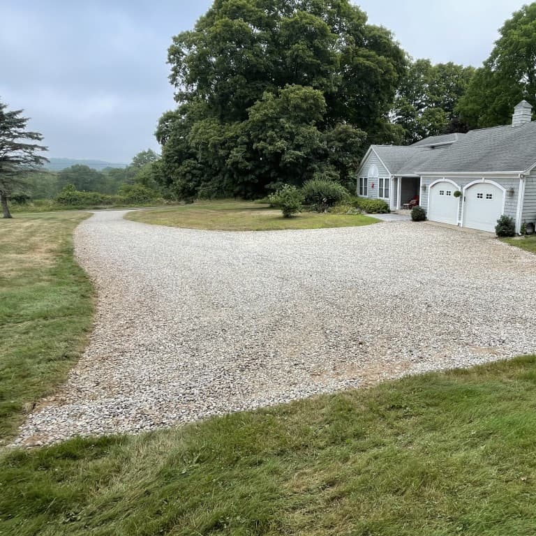 A gravel driveway curves toward a white house with a gray roof and double garage, showcasing landscape construction by Nyman Services, all surrounded by green grass and trees on a cloudy day.