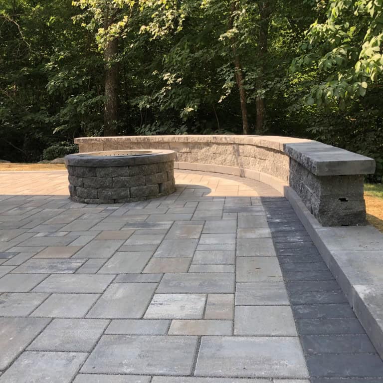 Circular stone fire pit and curved stone bench on a paved patio, expertly crafted by Nyman Services, showcase quality landscape construction and are surrounded by trees and greenery in the background.