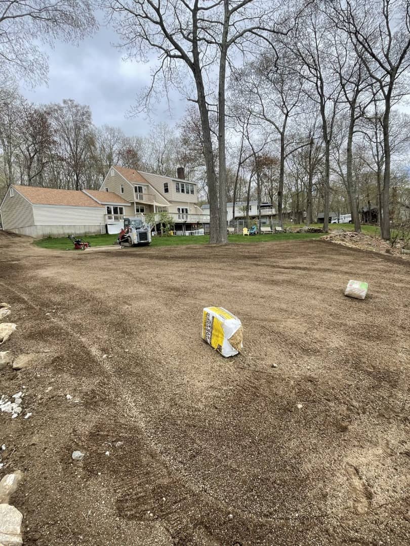 A yard with freshly leveled soil by Nyman Services, two bags of landscape construction material on the ground, a house and pickup truck in the background, and leafless trees surrounding the area.