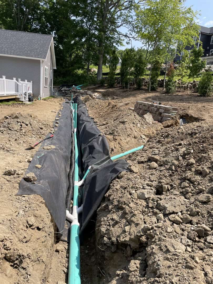 A trench with green PVC pipes and black fabric liner runs through a dirt yard next to a house, showcasing ongoing underground utility installation by Nyman Services, specialists in landscape construction.
