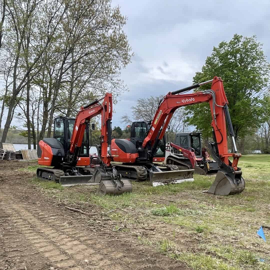 Three orange excavators from Nyman Services are parked on grass near trees, with a cloudy sky above and landscape construction materials visible in the background.