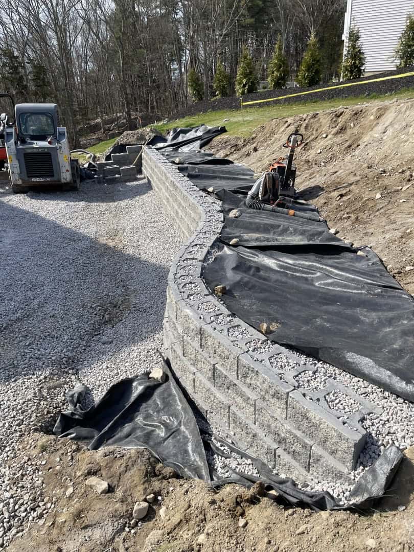 Curved retaining wall under construction with concrete blocks, gravel, and black geotextile fabric; landscape construction by Nyman Services, with construction equipment and trees in the background.