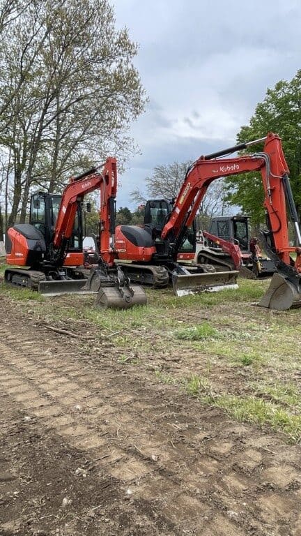 Three red excavators from Nyman Services are parked on a grassy, partially cleared area, ready for landscape construction, with trees in the background under a cloudy sky.