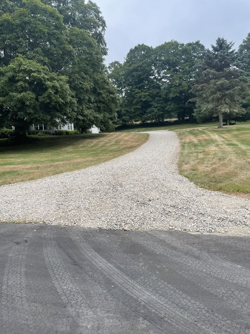 A gravel driveway curves through a grassy lawn with large trees in the background under an overcast sky, showcasing expert landscape construction by Nyman Services.