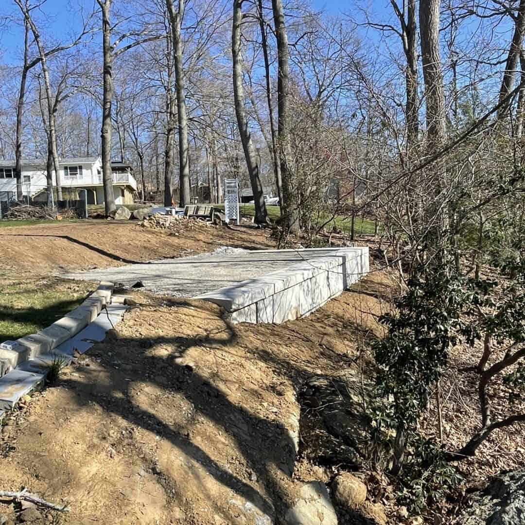 A recently built concrete retaining wall by Nyman Services runs along a dirt slope near trees, with expert landscape construction visible against houses and a clear blue sky in the background.