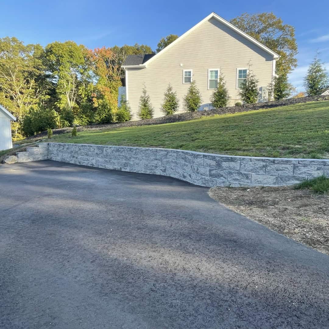 A paved driveway curves alongside a retaining wall made of stone blocks, showcasing expert landscape construction by Nyman Services, leading up to a beige house with a sloped yard and trees in the background.